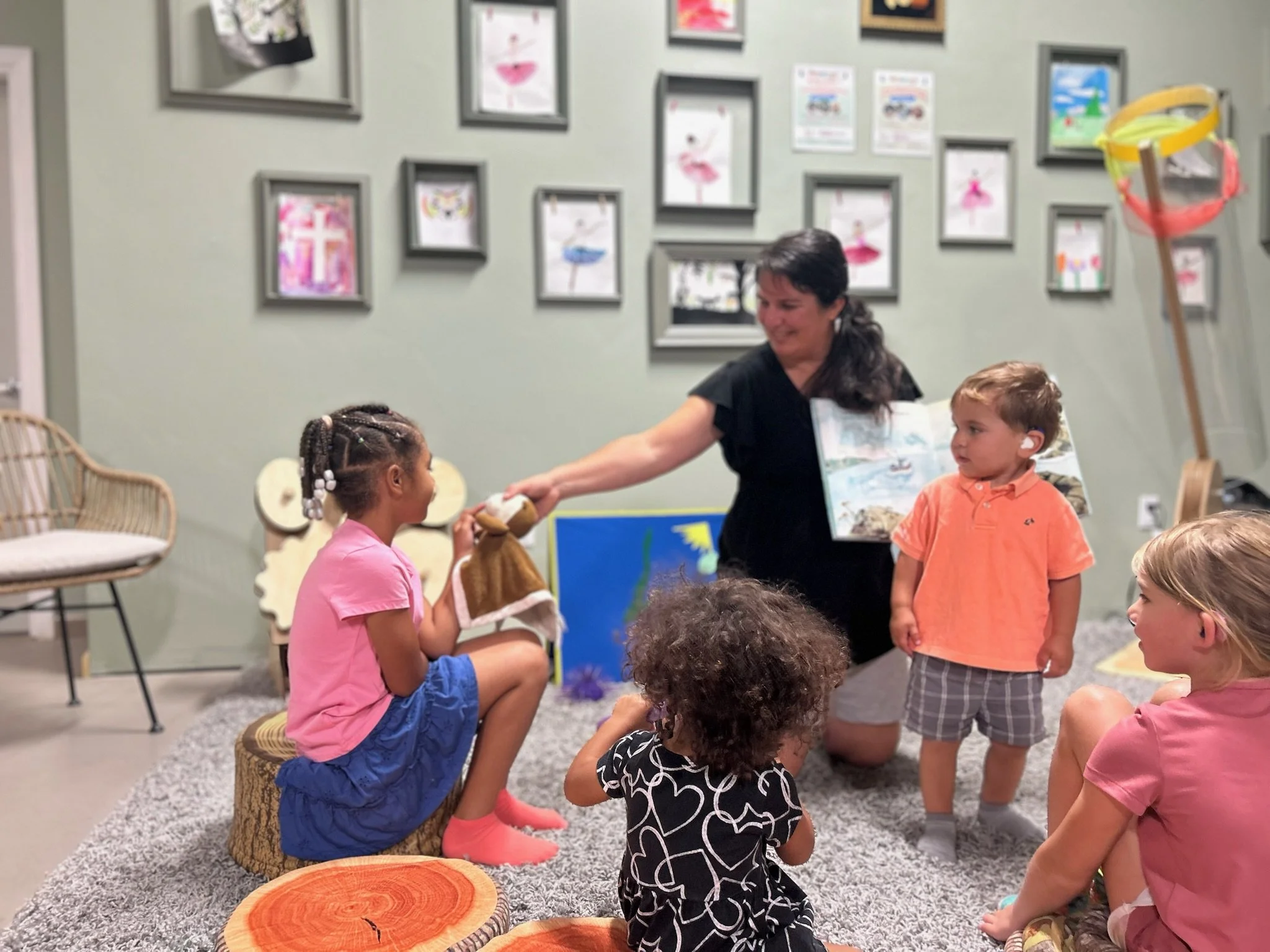 A woman leads a puppet show for four children in a colorful room decorated with framed artwork on the wall. The children are sitting on a gray carpet. The woman is holding a puppet and appears to be interacting with the children, who are watching her attentively.