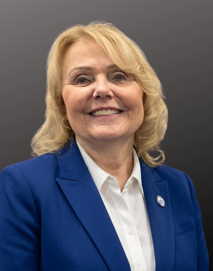 A smiling woman with blonde hair in a blue blazer and white shirt, standing against a gray background.