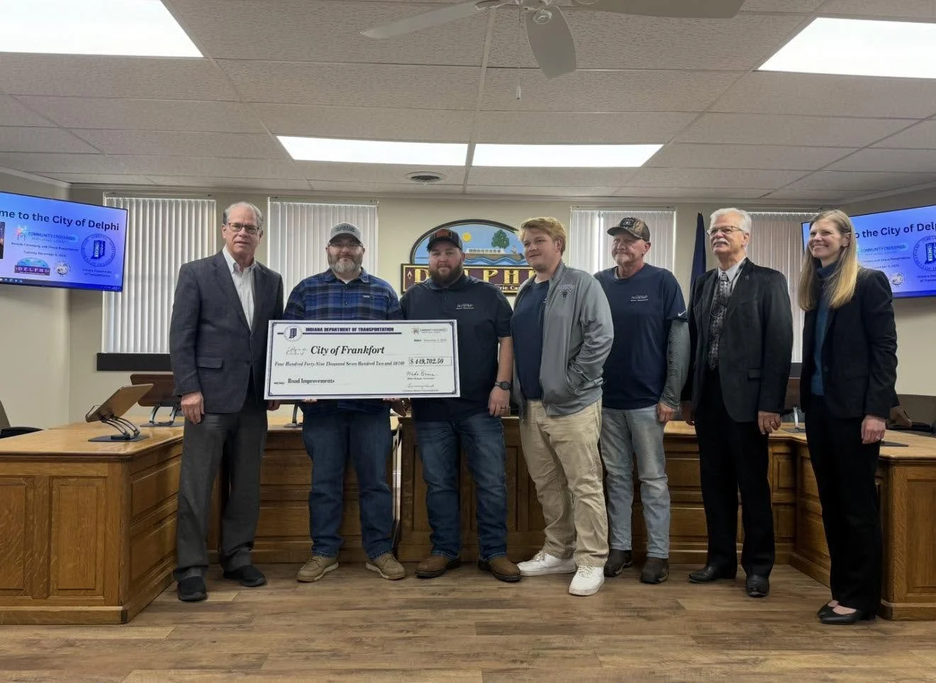 Group of seven people standing in a meeting room, holding a large check for road improvements issued to the City of Frankfort. Two people are holding the check, with others lined up behind them. Two screens behind display a message from the City of Delphi.