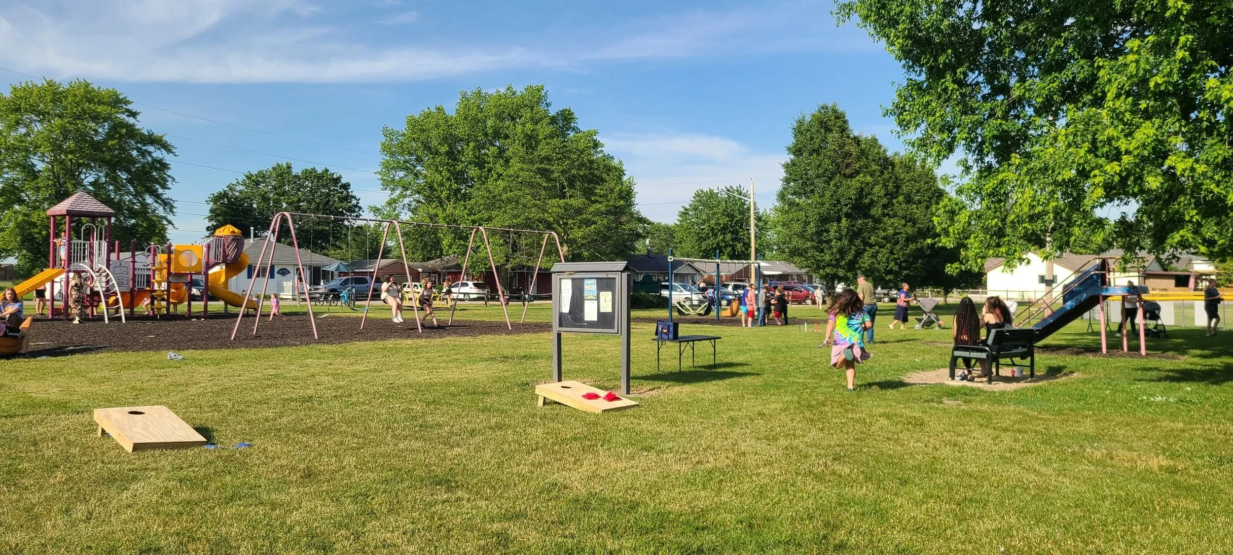 Children and adults enjoying a playground with swings, slides, and a climbing structure in a park on a sunny day.