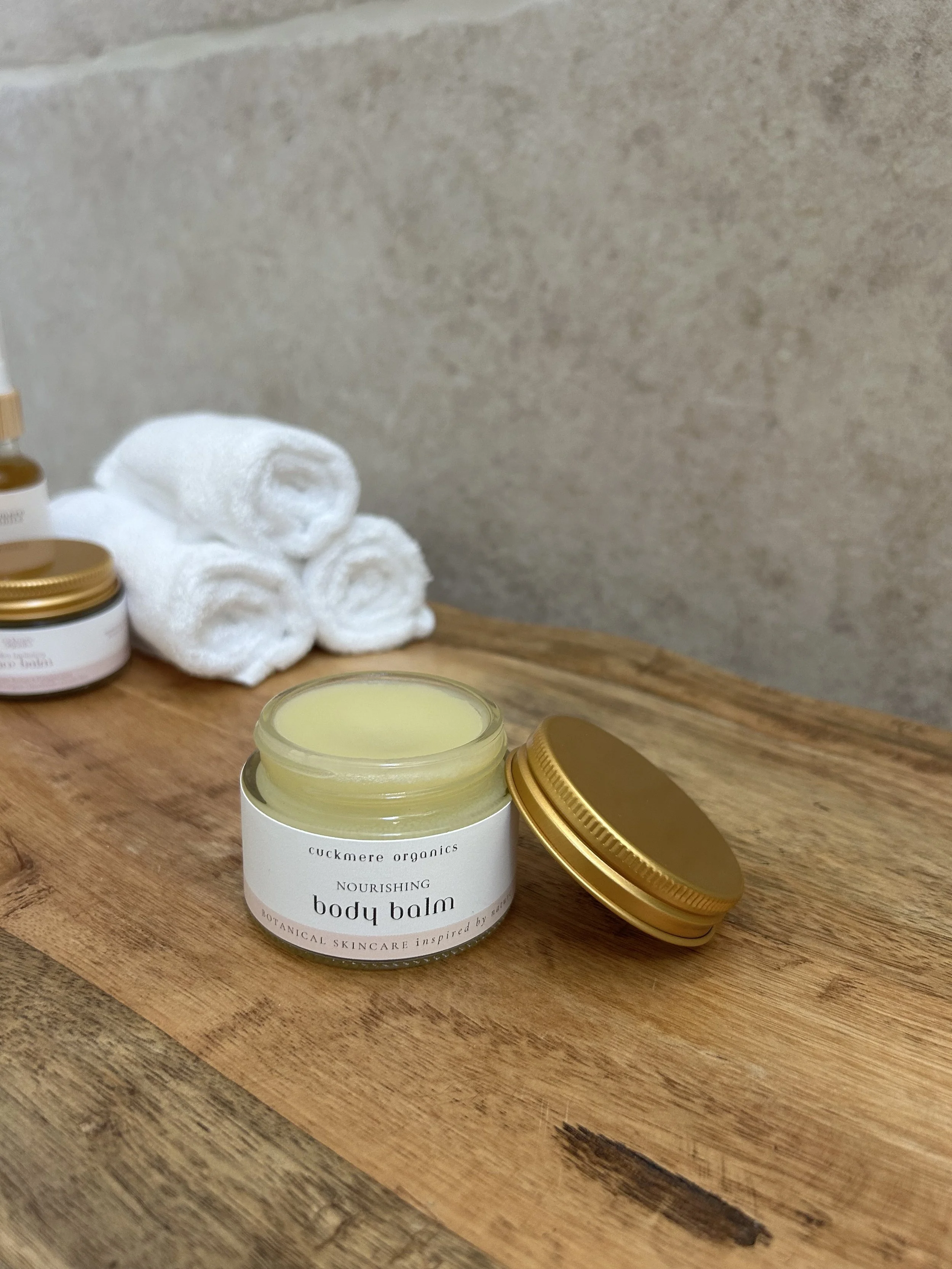 Cuckmere Organics Nourishing Body Balm in an open glass jar with a gold lid beside it, on a wooden surface with rolled white towels behind.
