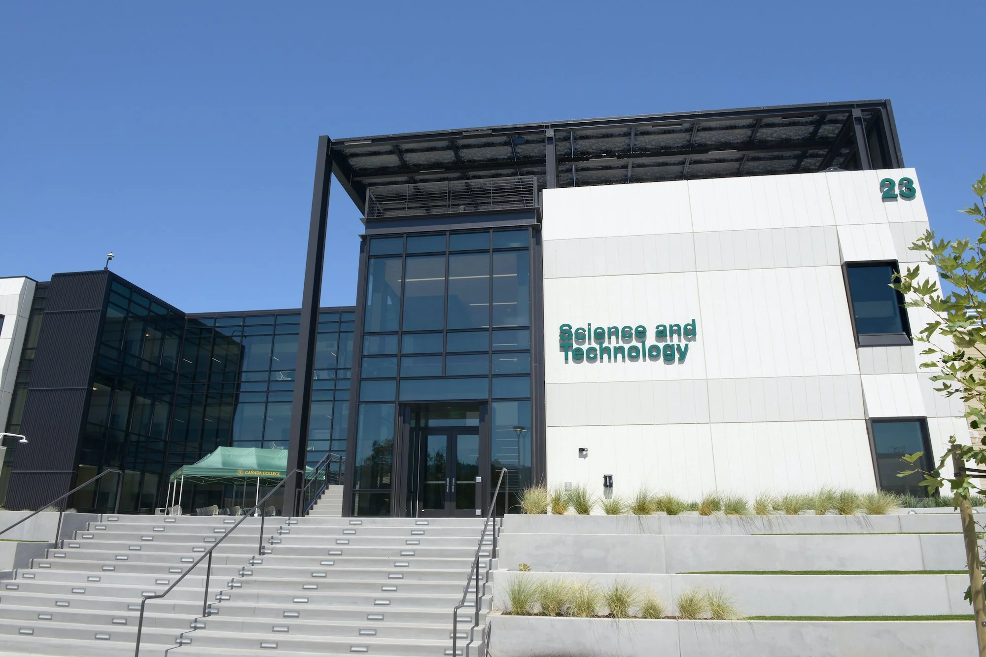 Modern educational building with large glass windows, stairs, and the words "Science and Technology" on the front, under a clear blue sky.