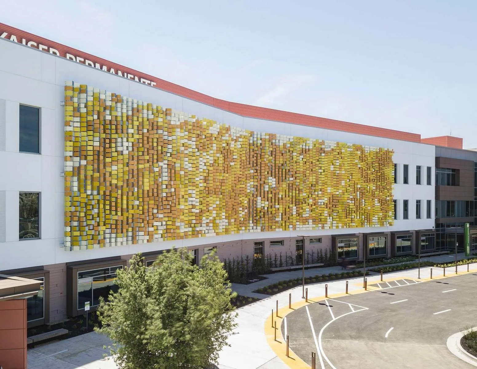 Modern hospital building with a colorful 3D tile mural on the upper wall, parking lot in the foreground, and trees lining the sidewalk.