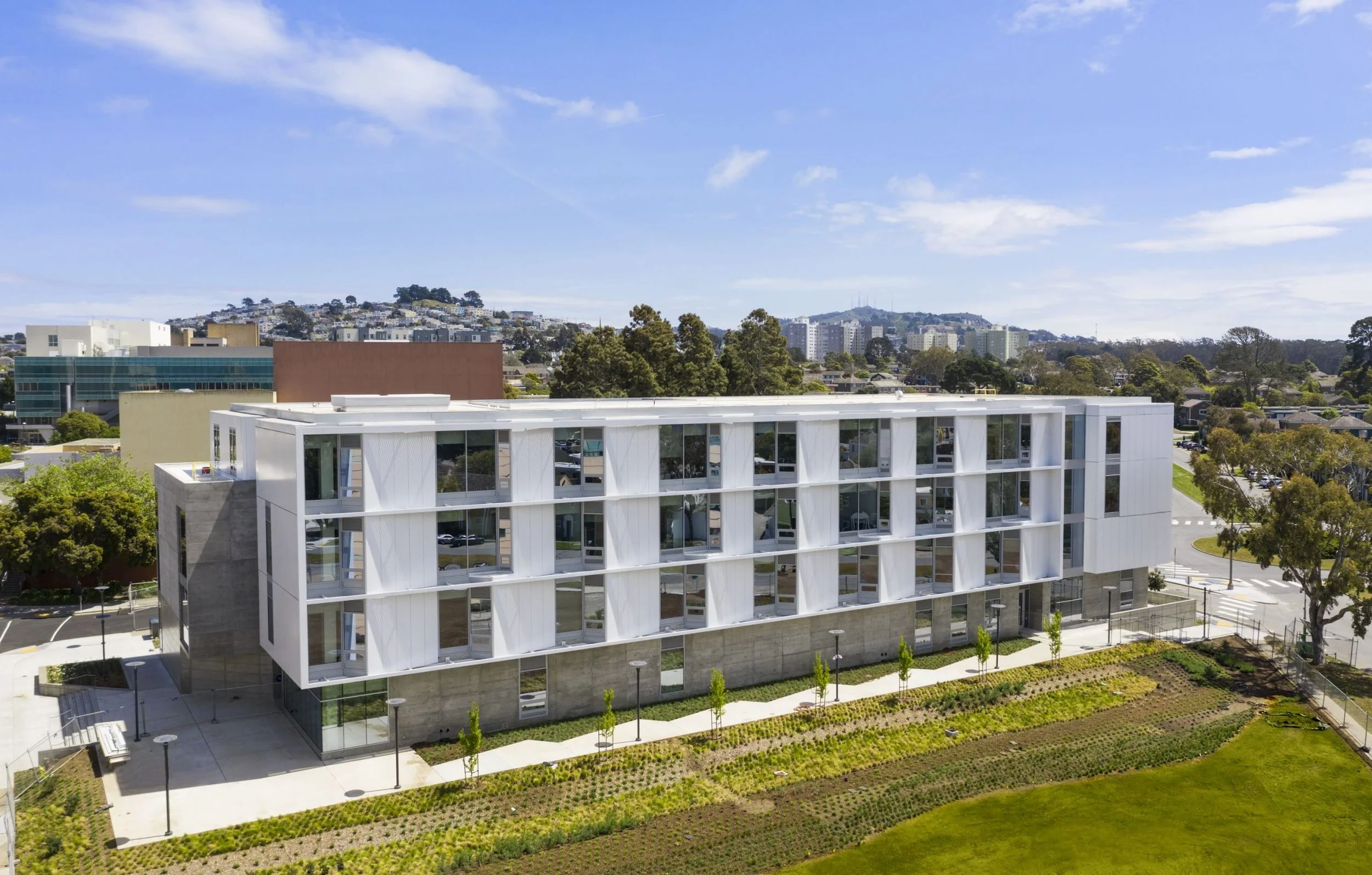 A modern white building with large glass windows surrounded by greenery and trees under a partly cloudy sky.