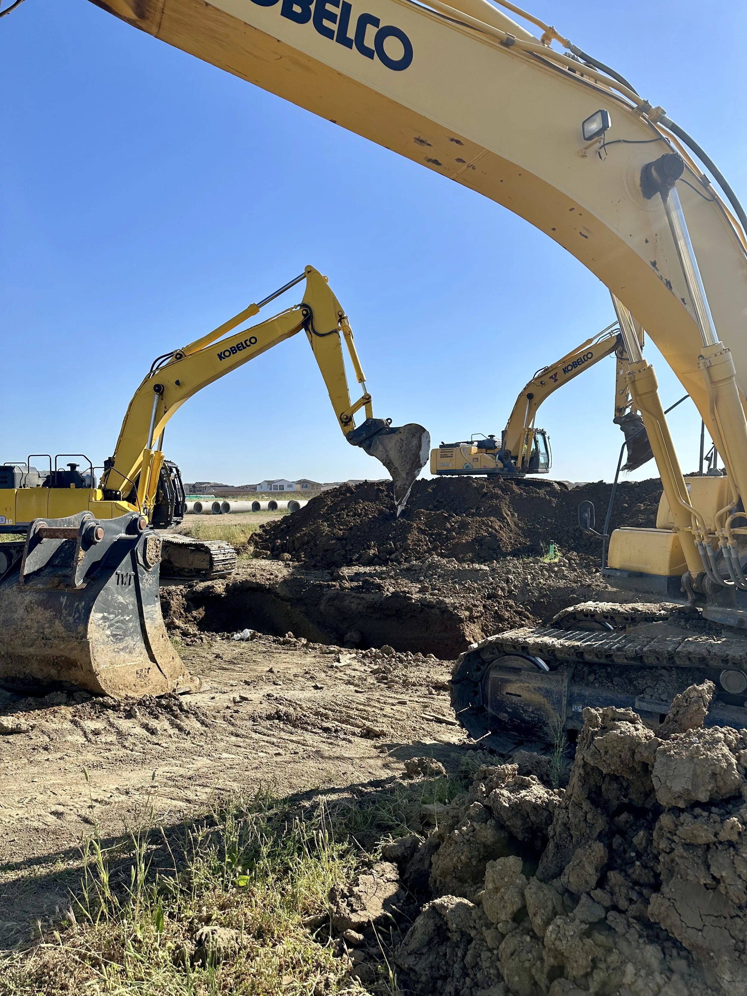 Construction site with multiple yellow excavators digging into the ground under a clear blue sky. Houses are visible in the background.