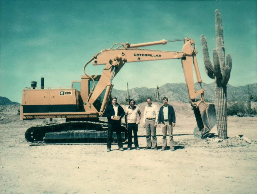 Four men standing in front of a large yellow Caterpillar excavator with a cactus in the desert landscape and mountains in the background.