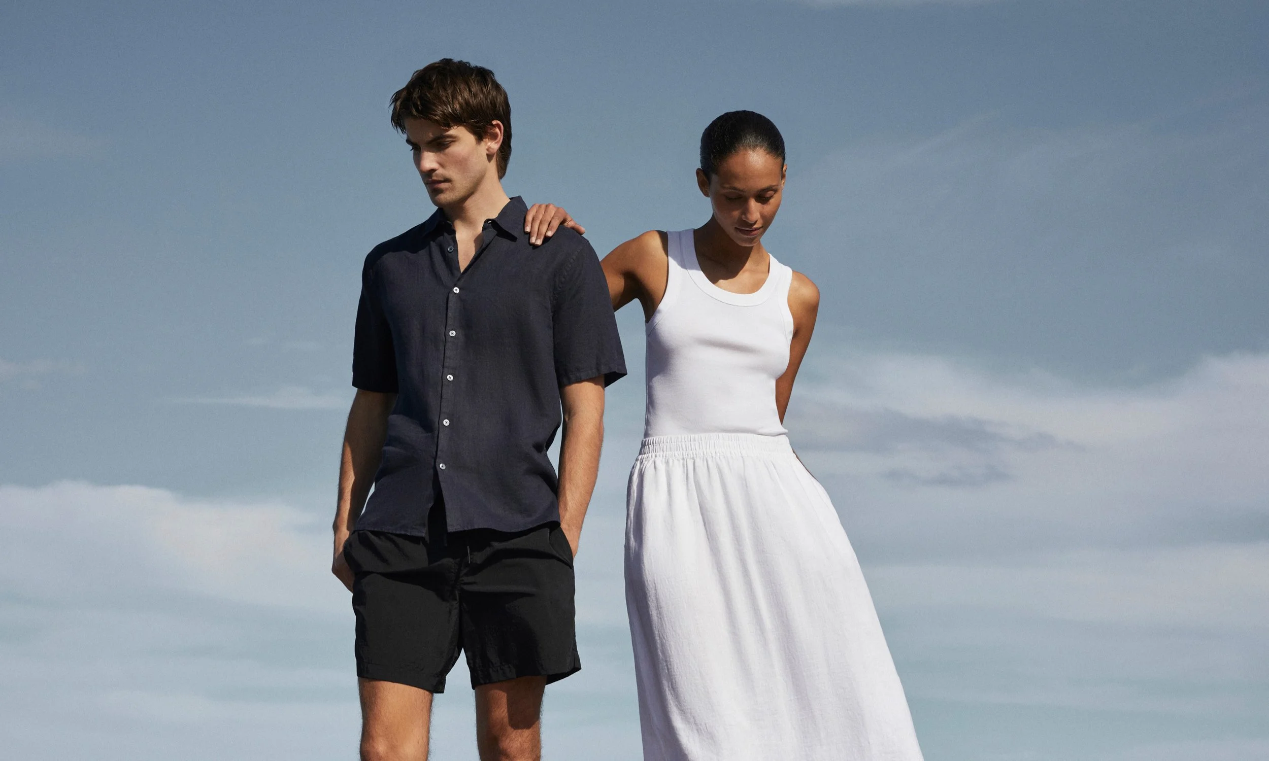 A young man and woman standing outdoors against a cloudy sky, with the woman placing her hand on the man's shoulder.