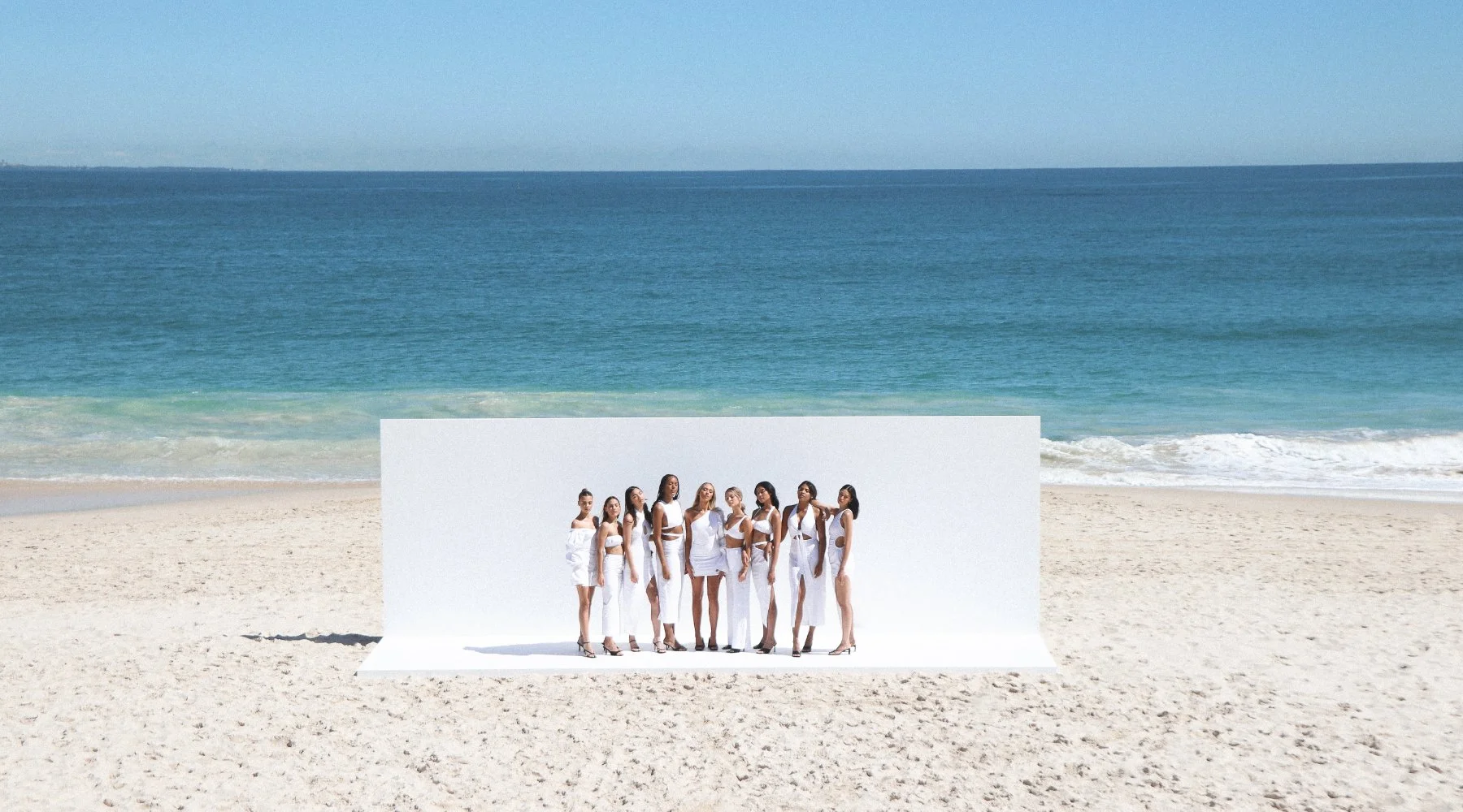 A group of women in white clothing standing on a white platform on a sandy beach near the ocean.