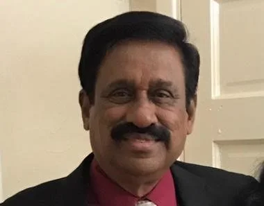 A man with short, dark, slightly wavy hair, a short beard, and mustache, wearing a white dress shirt with black polka dots, standing against a plain white wall.