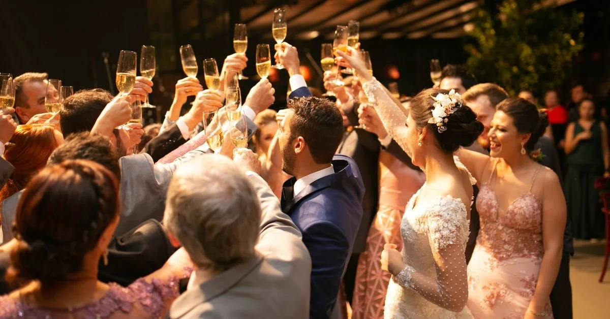 A group of people celebrating at a wedding reception, raising glasses of champagne for a toast, with a bride and groom in the center, surrounded by friends and family.