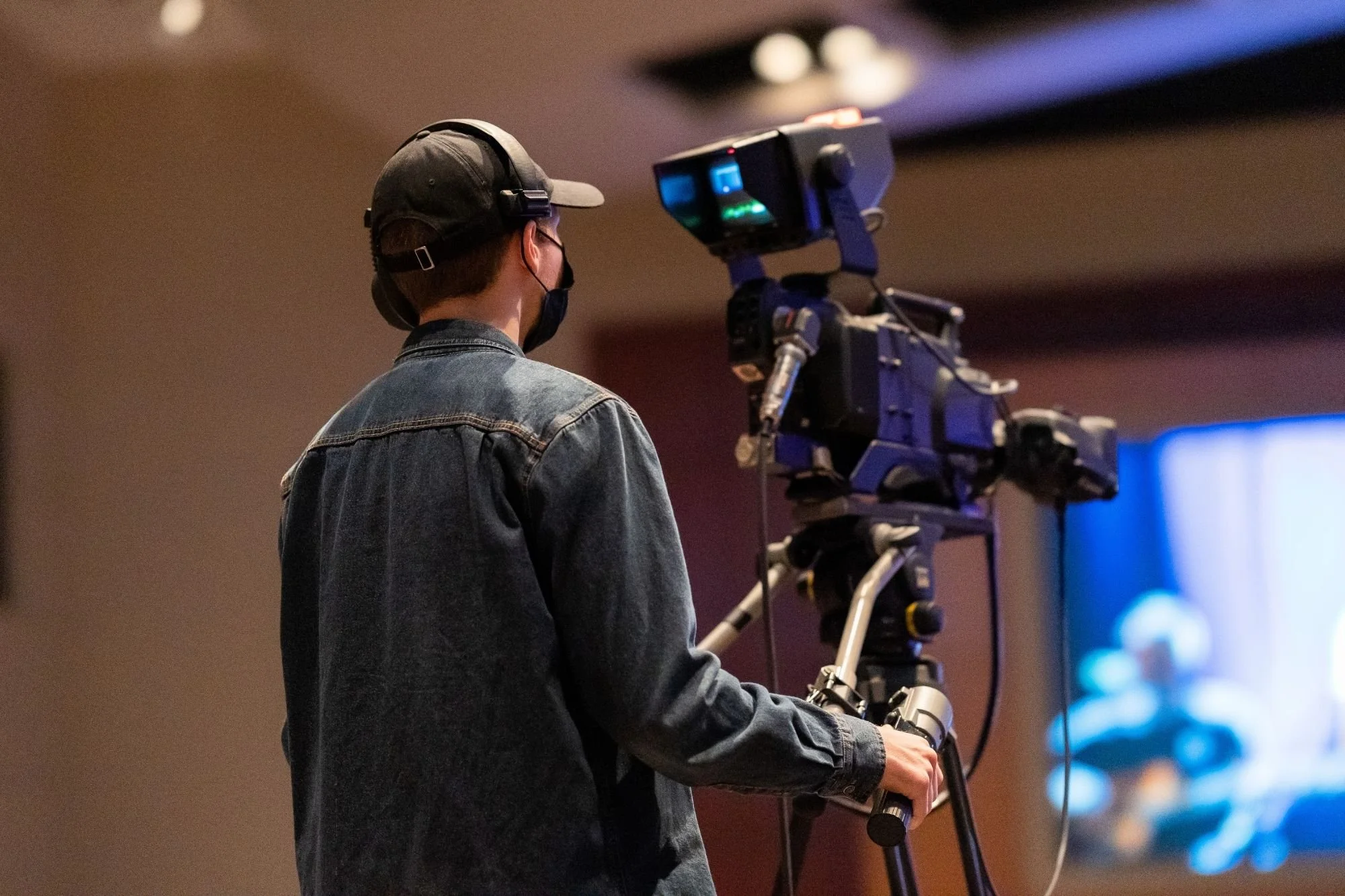 videographer operating broadcast camera on tripod at indoor venue