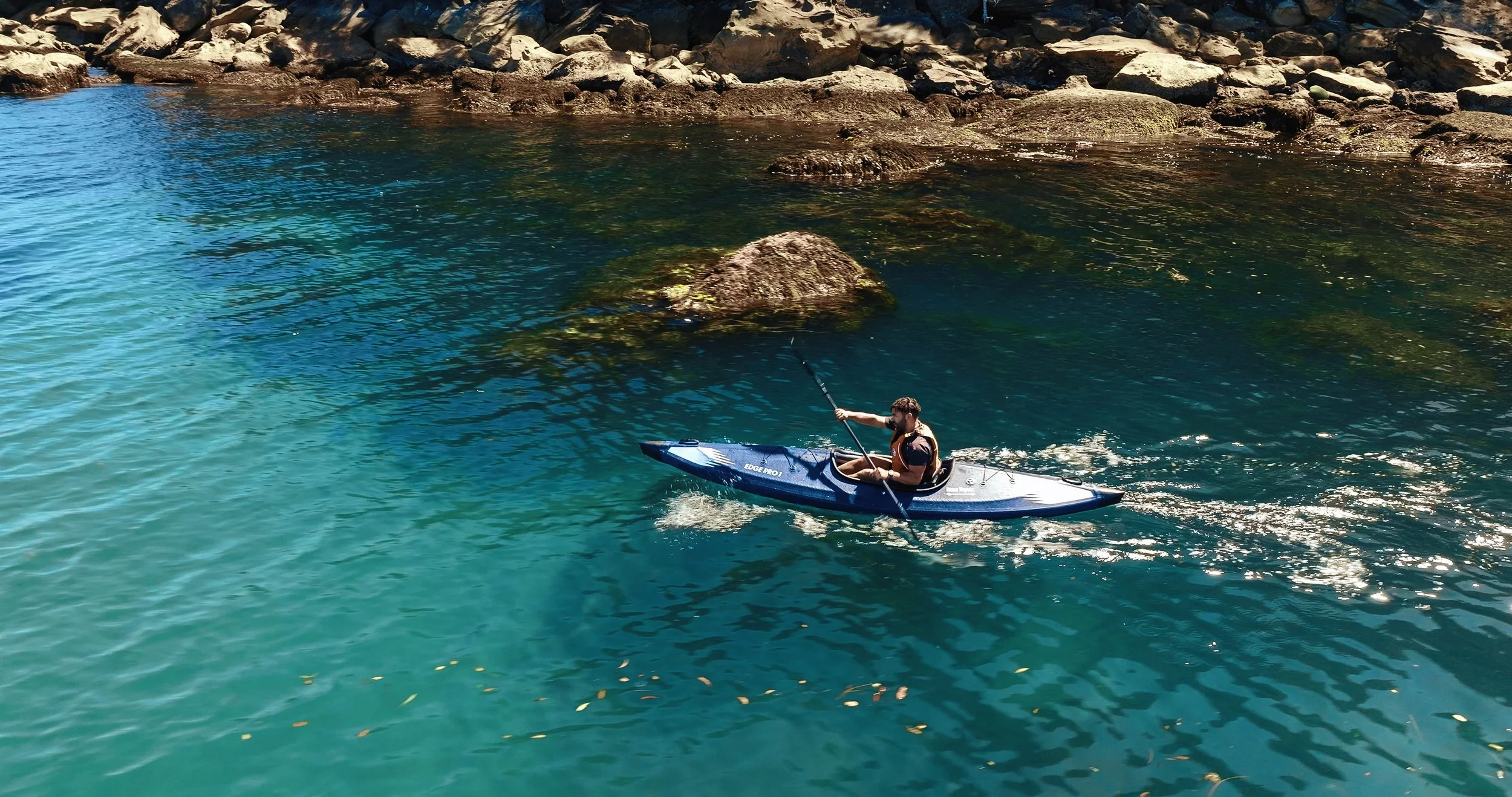 Person paddling a blue kayak on clear blue water near rocky shoreline