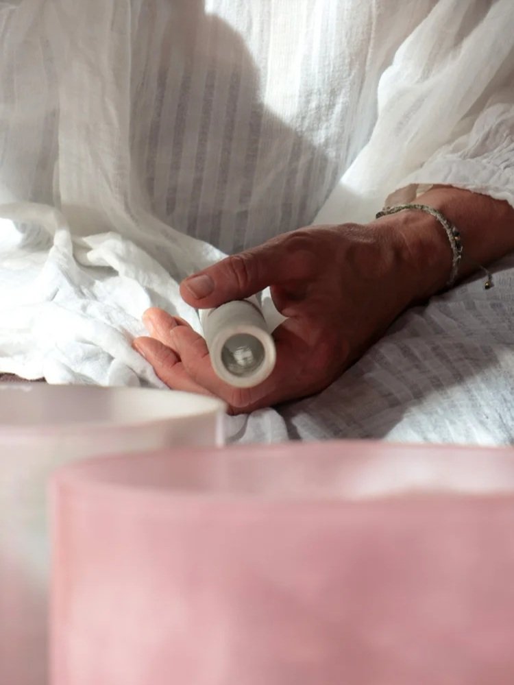 Close-up of an elderly person's hand holding a small white object, possibly a medicine container, resting on white fabric or clothing, with a pink and white blurry object in the foreground.