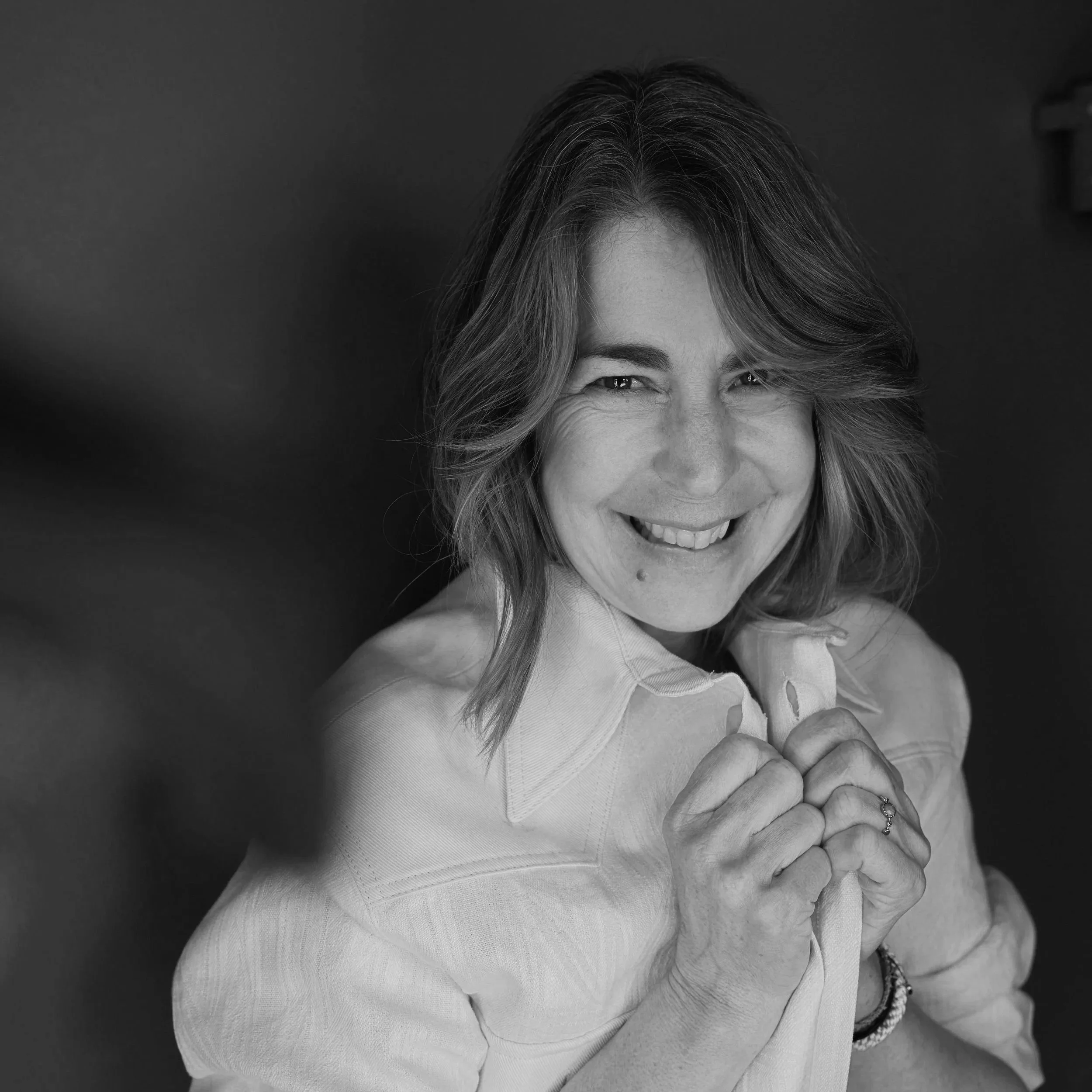 A black and white portrait of a smiling woman with shoulder-length hair, holding her shirt collar with both hands, looking directly at the camera.