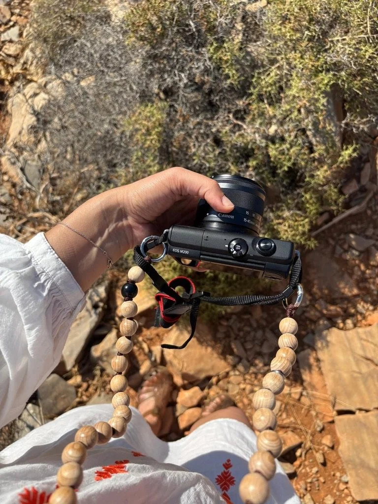 A person holding a Canon EOS M200 camera with a beaded strap, standing on rocky ground with dry foliage, dressed in white clothing.