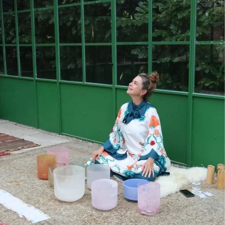 Person seated on a mat surrounded by singing bowls on a mandala-patterned wooden floor.