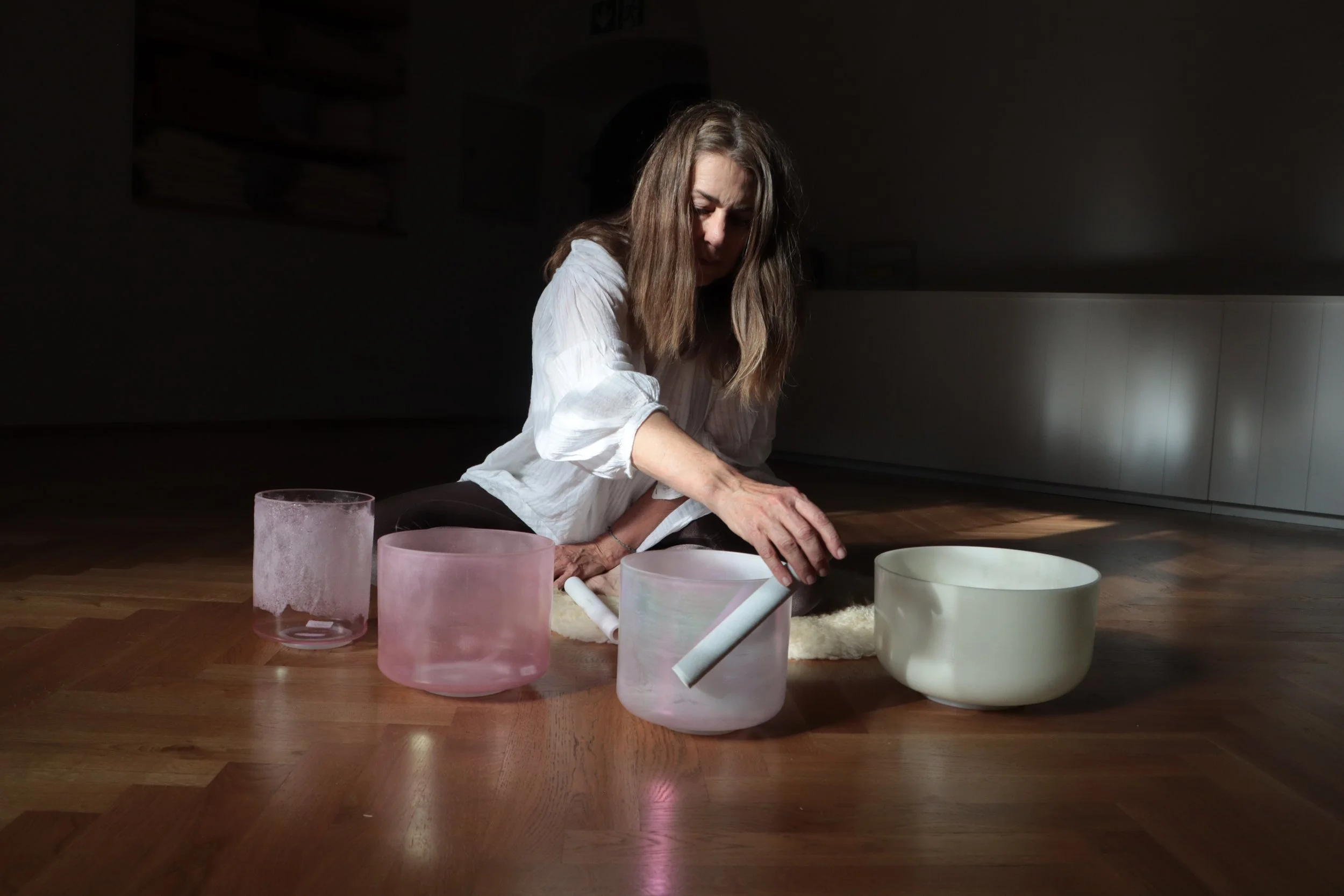 A woman with dark hair in a bun sits on a patterned rug, looking at colorful glass singing bowls arranged on the floor in front of her.