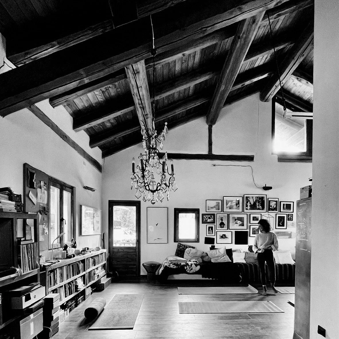 Black and white photo of a cozy living room with wooden beams and a chandelier. The room features a sofa, bookshelves, yoga mats on the floor, and framed pictures on the wall. A person stands near the wall.