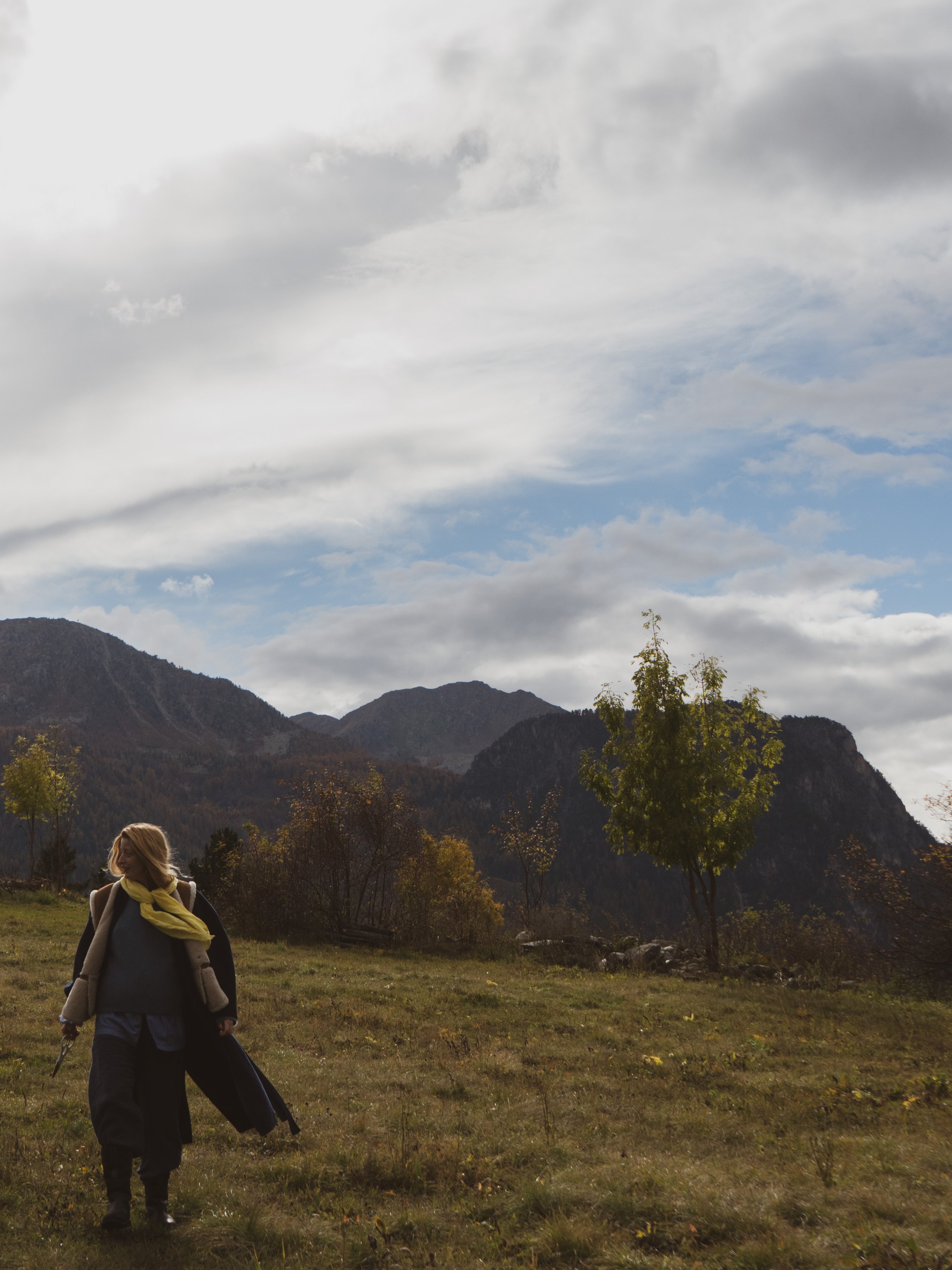 A woman with blond hair, wearing a dark jacket, blue pants, and a yellow scarf, walking in a grassy field with trees and mountains in the background under a cloudy sky.
