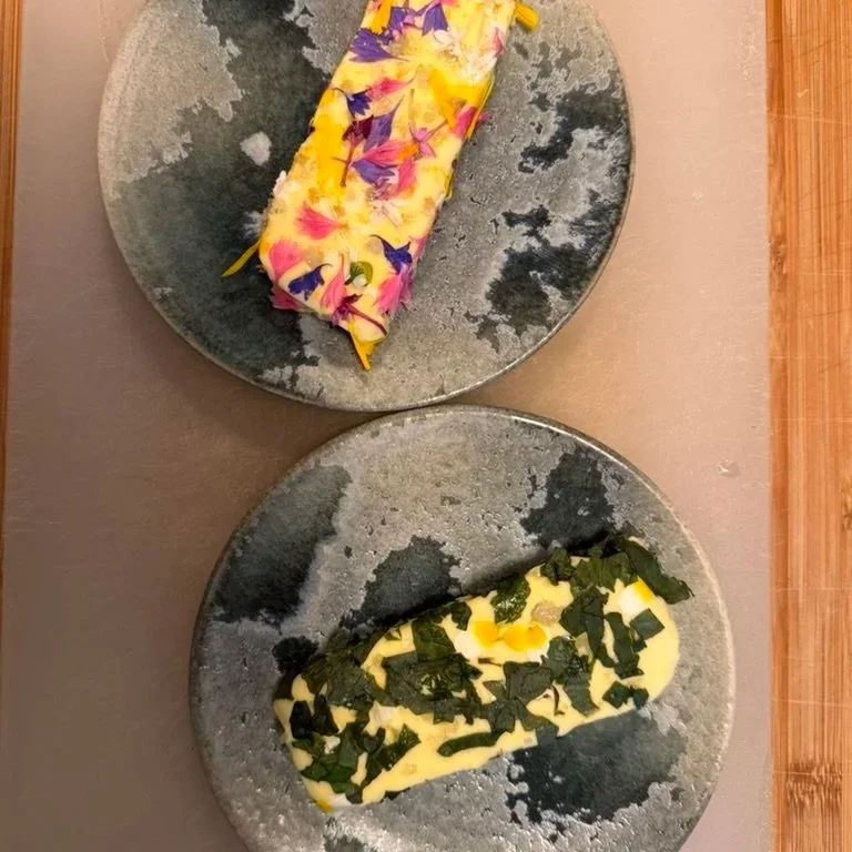 Two plates each with a piece of sushi, one topped with colorful edible flower petals and the other with chopped green herbs on a gray ceramic plate.