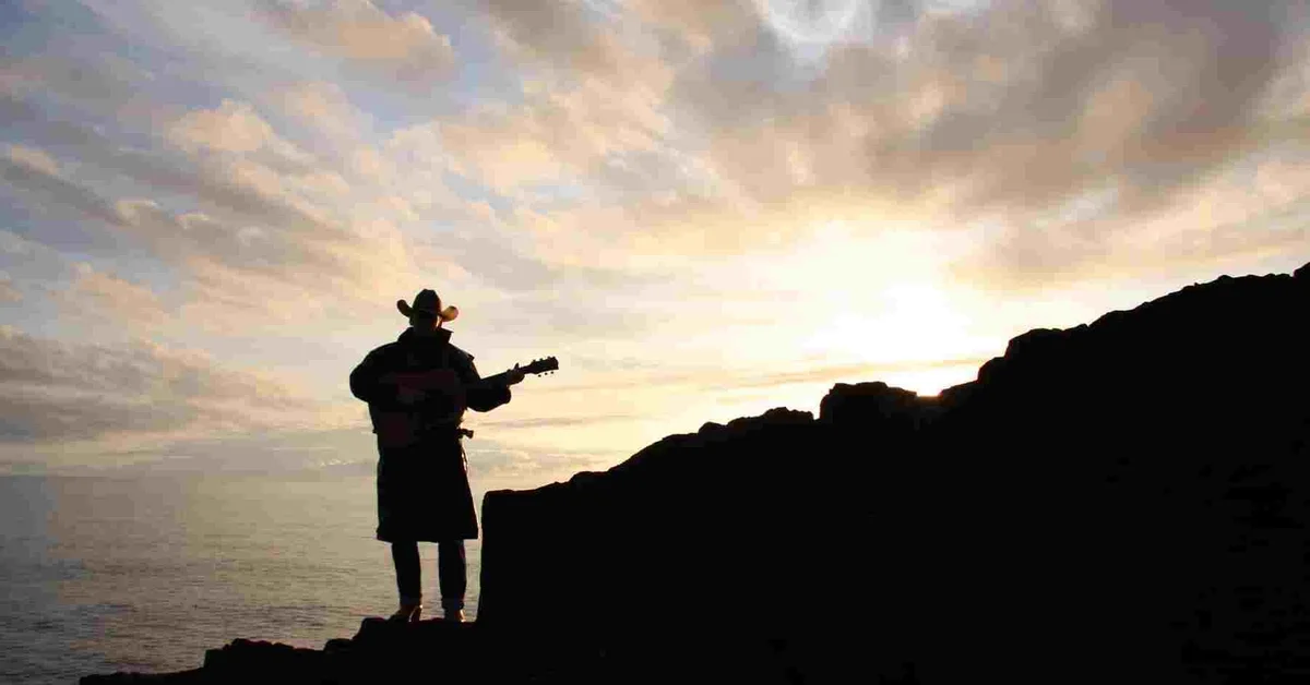 Silhouette of a person wearing a cowboy hat, standing on rocks near the ocean, playing a guitar at sunset with a cloudy sky.