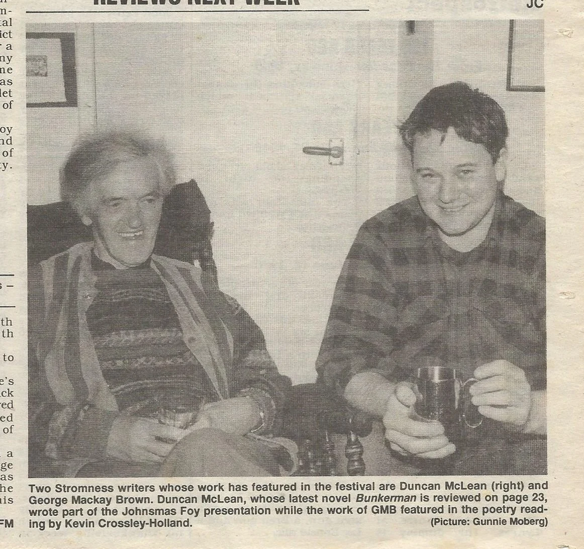 A black and white photo of two authors, a woman and a man, sitting indoors. The woman, on the left, is smiling and wearing a patterned sweater with a scarf. The man, on the right, is also smiling, wearing a plaid shirt, and holding a coffee mug.