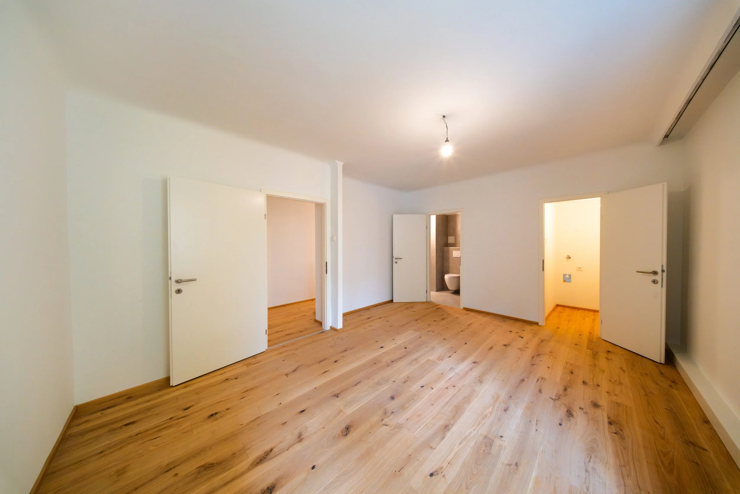 Leerer Raum mit weißen Wänden, Holzfußboden, drei offene Türen, eine führt zu einem Badezimmer, eine andere zu einem Abstellraum, und eine dritte zu einem anderen Raum, Deckenleuchte ohne Abdeckung.