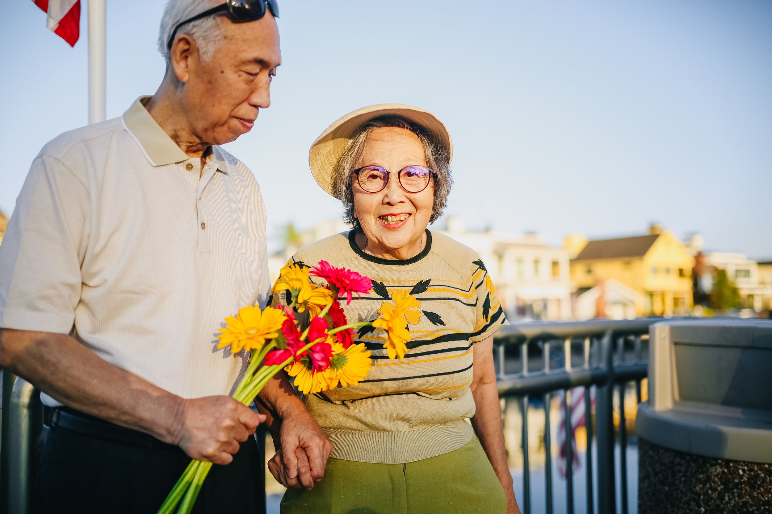 A sweet asian couple where the man is holding a bouquet of flowers for his wife; they are both holding hands. It appears to be in the golden hour at sunset in the evening.