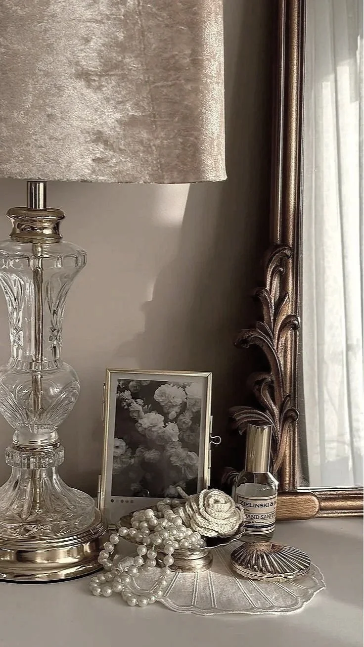 Decorative table top with a glass lamp with a large beige lampshade, a framed black-and-white floral photo, a string of pearls, a shell, a small bottle of hand sanitizer, and a large decorative mirror with a wooden frame.