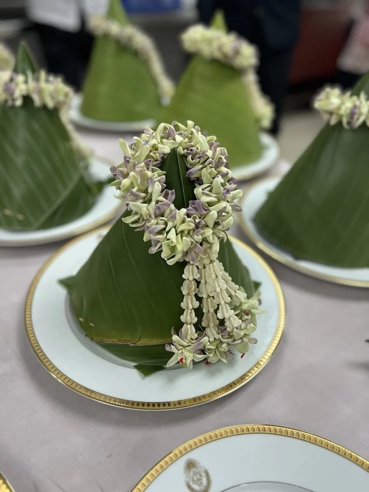 Conical green base wrapped in banana leaves with a decorative flower garland draped over it, placed on a fine china plate with a gold rim