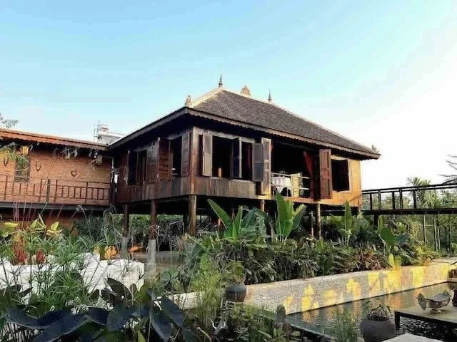 Traditional wooden house with a tiled roof, elevated on stilts, surrounded by lush green plants, under a clear blue sky.