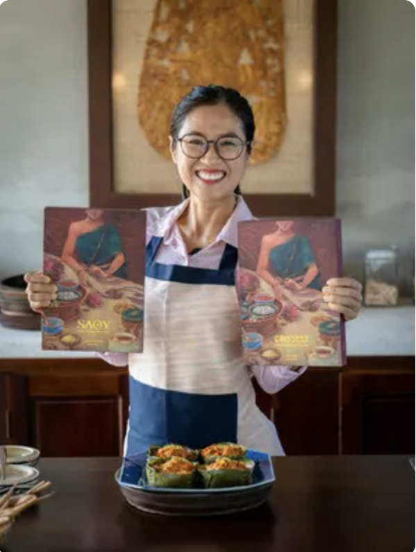 A woman smiling and wearing glasses, holding two magazines with a woman in a turquoise dress on the cover. There are four cupcakes on a plate in front of her on the table, and a few bowls and utensils are visible nearby. The background features a decorative wall hanging behind her.