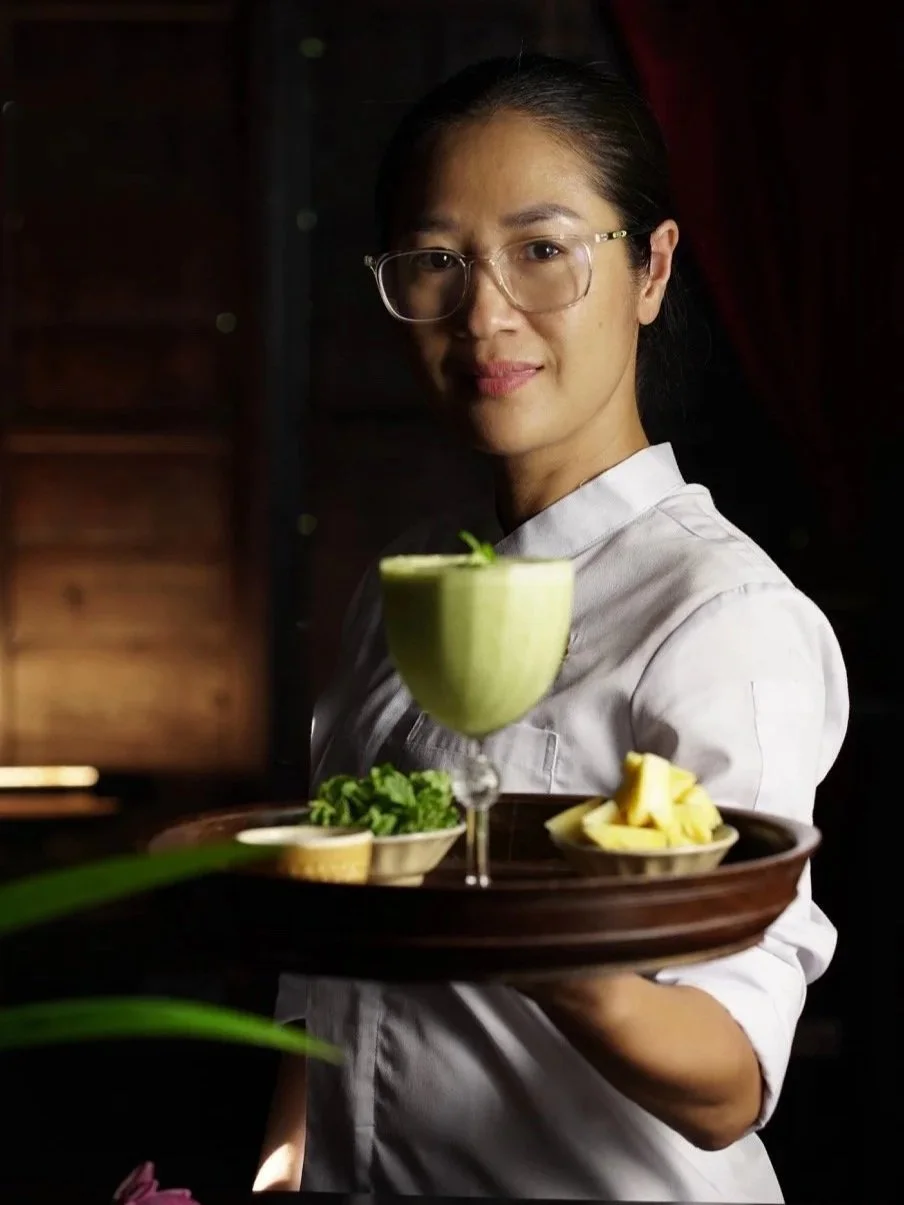 A woman with glasses and a white shirt holding a tray with a green beverage, lemon slices, and herbs, in a dimly lit setting.