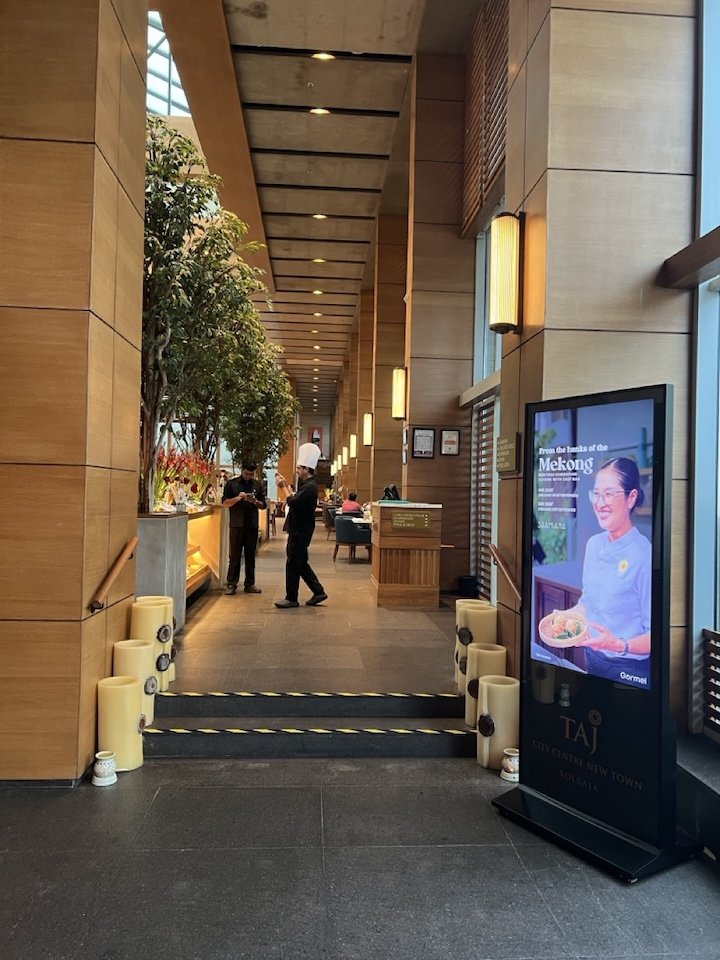 Interior of a modern restaurant or hotel lobby with wooden paneling, indoor plants, digital display, and staff members preparing or taking orders.