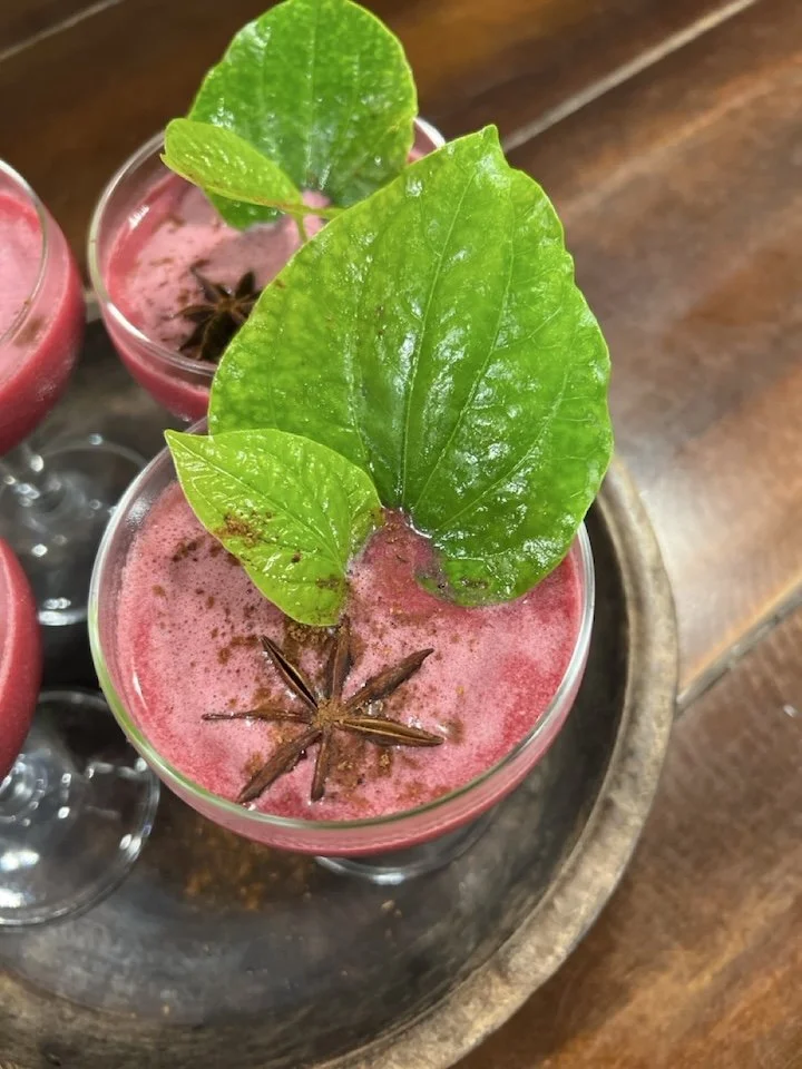 A pink frothy beverage in a glass garnished with large green leaves and star anise on top, placed on a wooden tray.