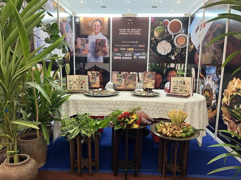 A booth with a white tablecloth displaying traditional food and illustrated books, surrounded by plants, with large posters and a banner promoting heritage and cultural content in the background.