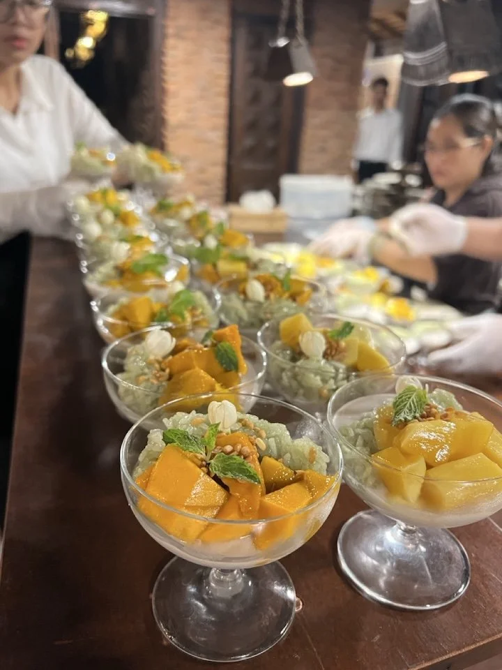 Multiple dessert cups filled with diced mango, some garnished with mint leaves and white decorative toppings, lined up on a wooden table in a restaurant setting.