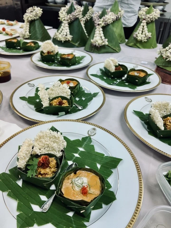 Traditional Indonesian dish called Nasi Tumpeng, featuring yellow rice shaped into a cone, surrounded by various side dishes served in banana leaf containers, on a white tablecloth with decorative floral carvings and green leaves.
