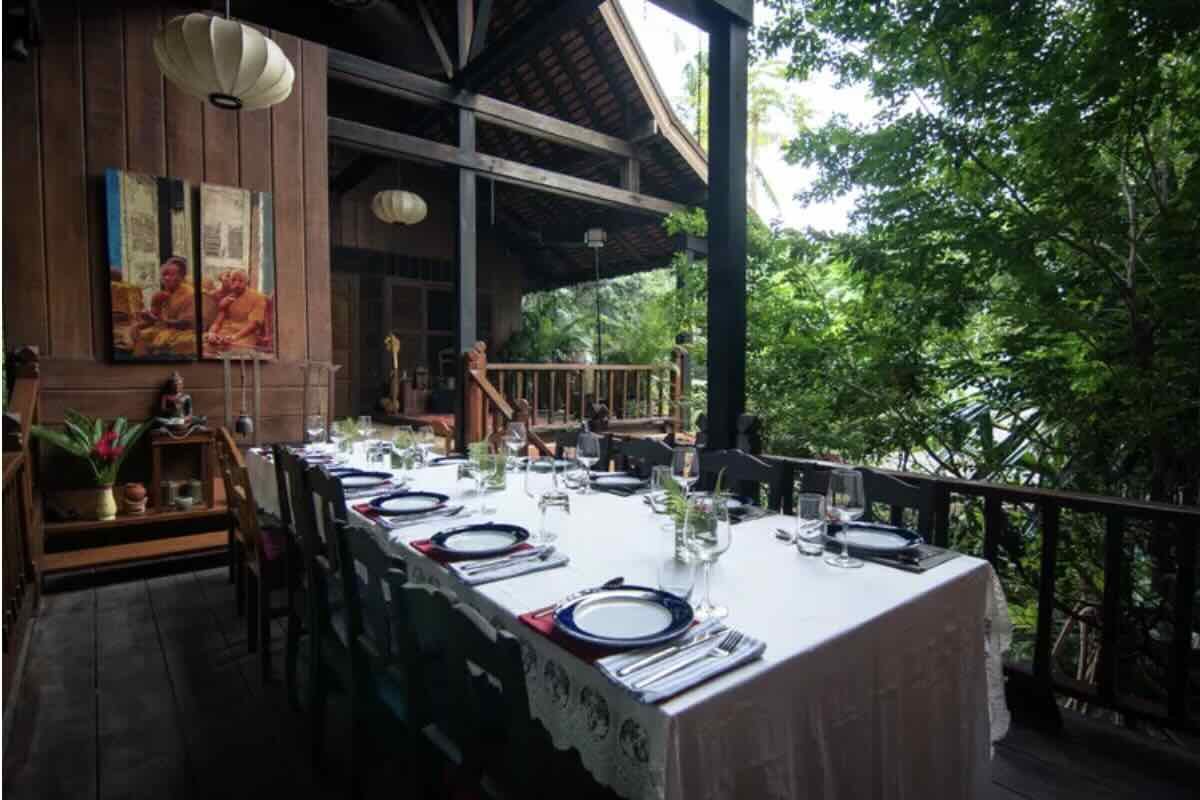 Outdoor dining table set with plates, glasses, and silverware on a white tablecloth, surrounded by lush green trees and a wooden porch.
