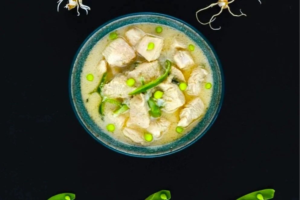 A bowl of creamy chicken curry with chicken pieces and sliced green chili peppers on top, placed on a black surface with some green chili slices and white flower-like garnishes around it.
