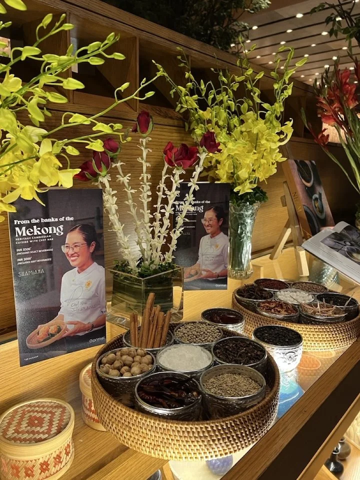 A display of spices and herbs in small jars on a wooden shelf, with fresh flowers in vases in the background and promotional posters featuring a smiling woman in white chef's attire, in an indoor setting.