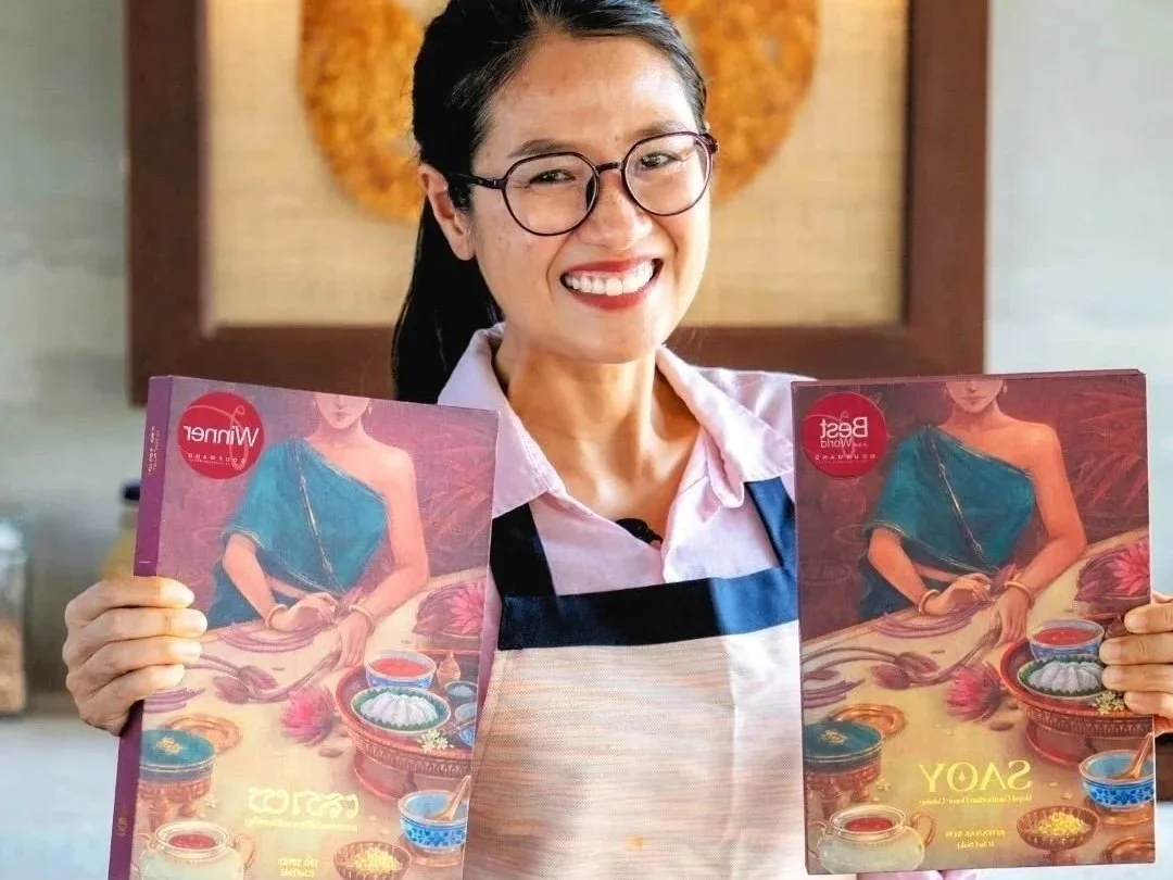 A woman with black hair, glasses, and a pink shirt smiling and holding two magazines with the cover art of a woman in a turquoise dress sitting at a table with food and drinks.