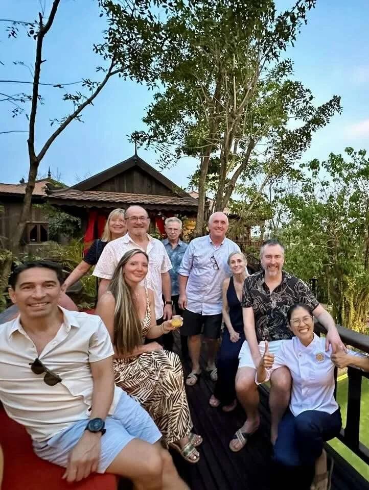 Group of people enjoying an outdoor gathering on a wooden deck with trees and a house in the background.