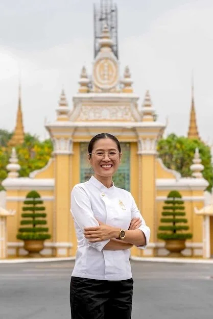 A smiling woman in a white chef's uniform standing outdoors in front of a colorful, ornate gate.