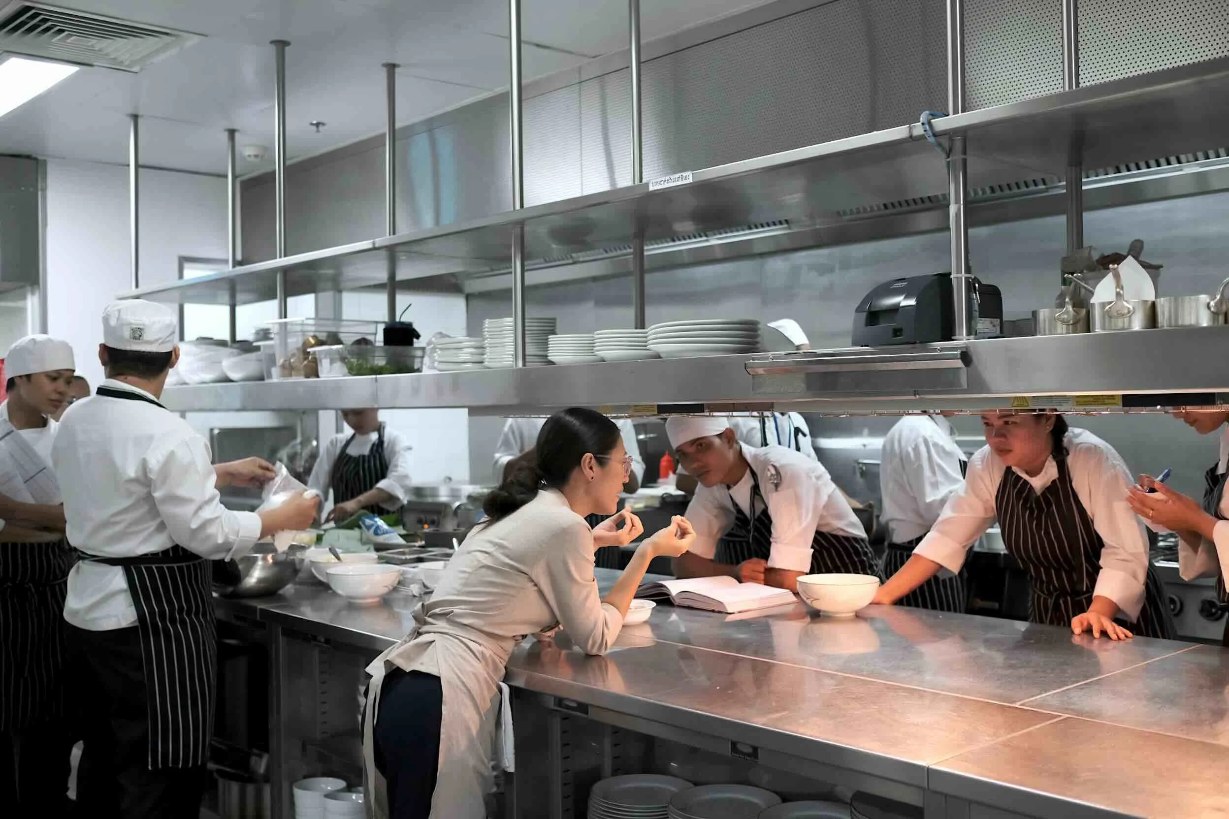 Chef Nak instructed Rosewood Hotel kitchen team as they're working together in a hotel kitchen with stainless steel counters, plates, and cooking equipment