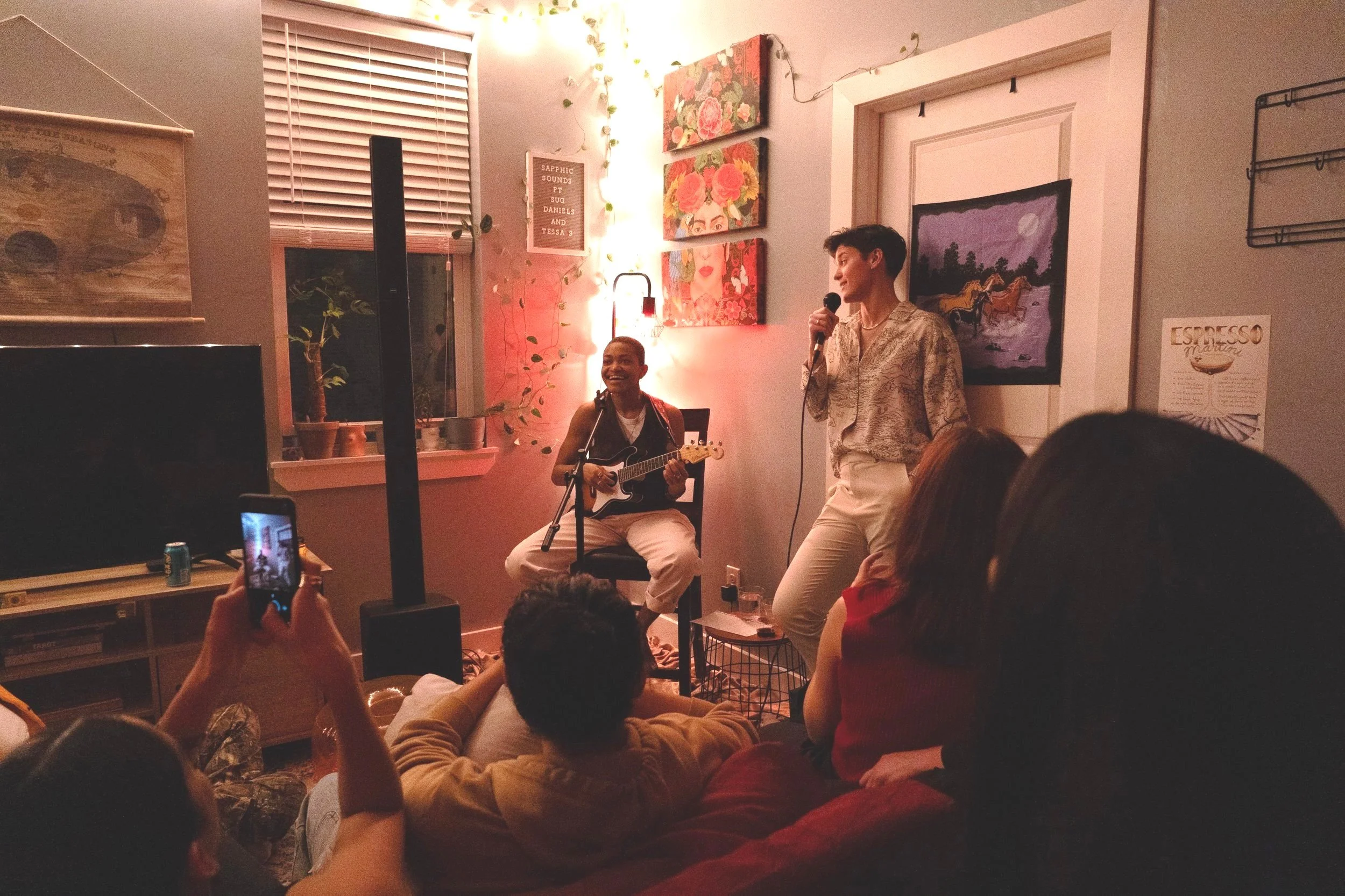 Two performers sing and play guitar in a cozy, decorated room while a small audience watches and takes photos.