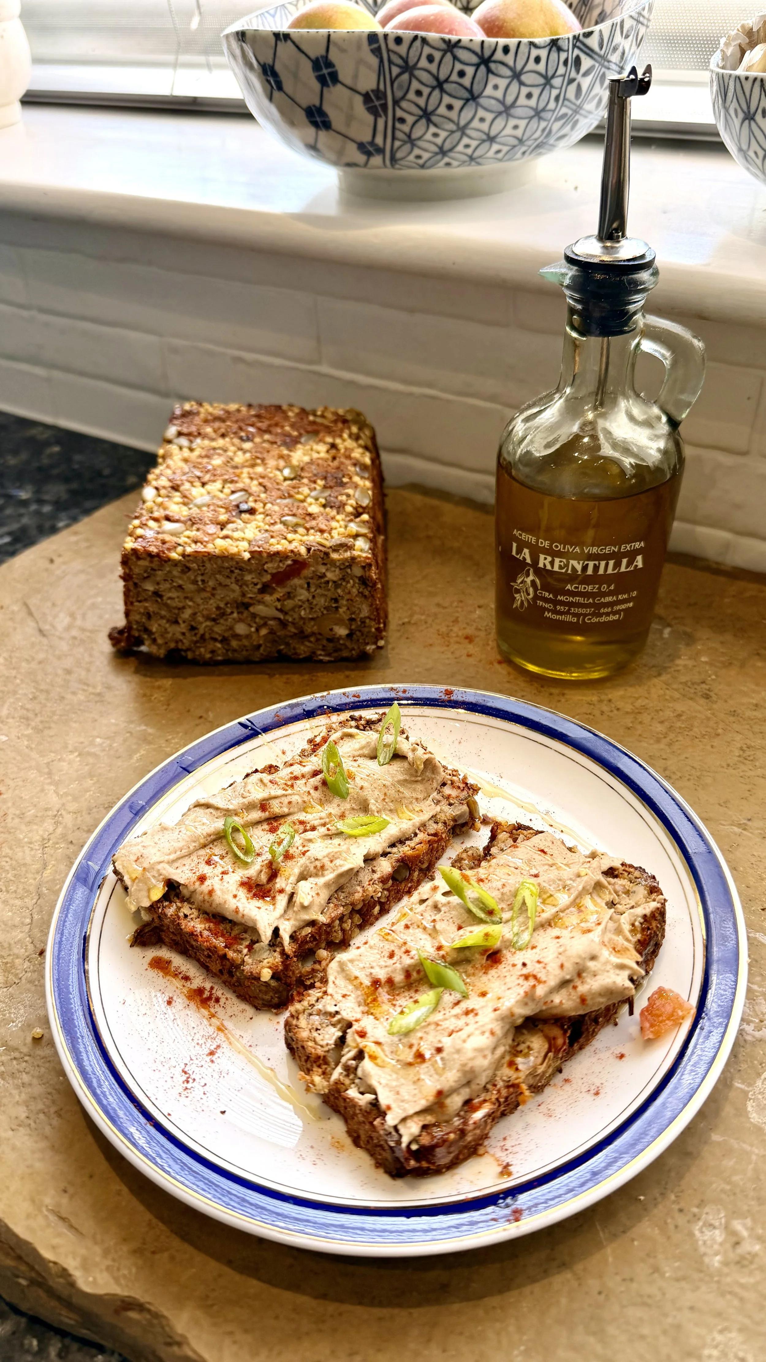 Two thick slices of homemade vegan seed and vegetable loaf topped with a creamy spread, spring onions, paprika and a drizzle of olive oil, served on a blue-rimmed plate. The remaining loaf and a bottle of extra virgin olive oil sit behind.