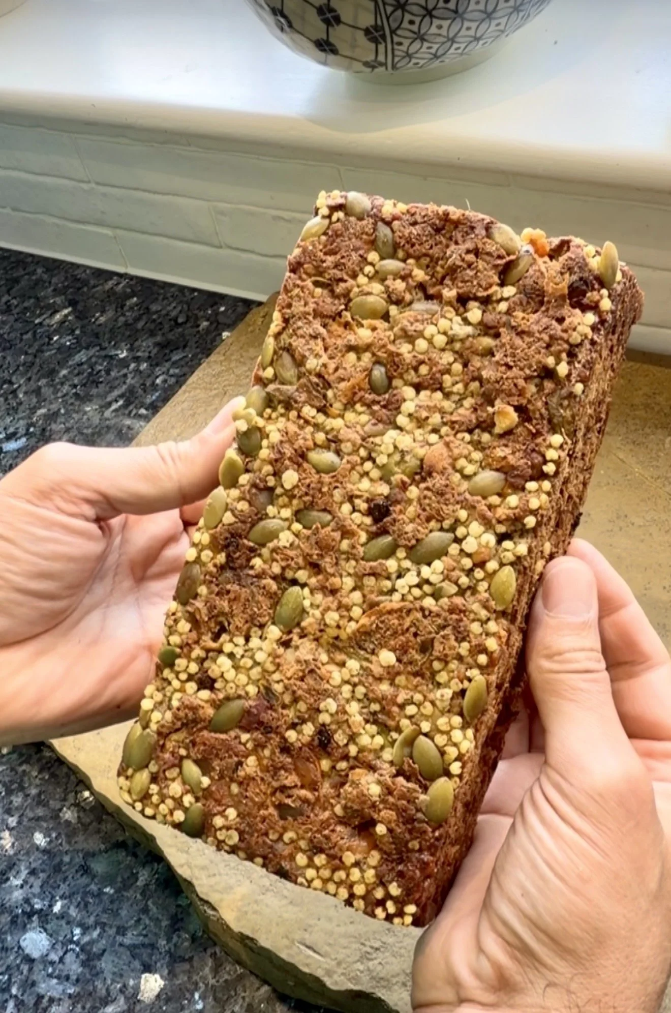 Hands holding a dense homemade vegan seed and vegetable loaf, showing its deeply browned crust studded with pumpkin seeds and small grains, against a kitchen worktop and window background.