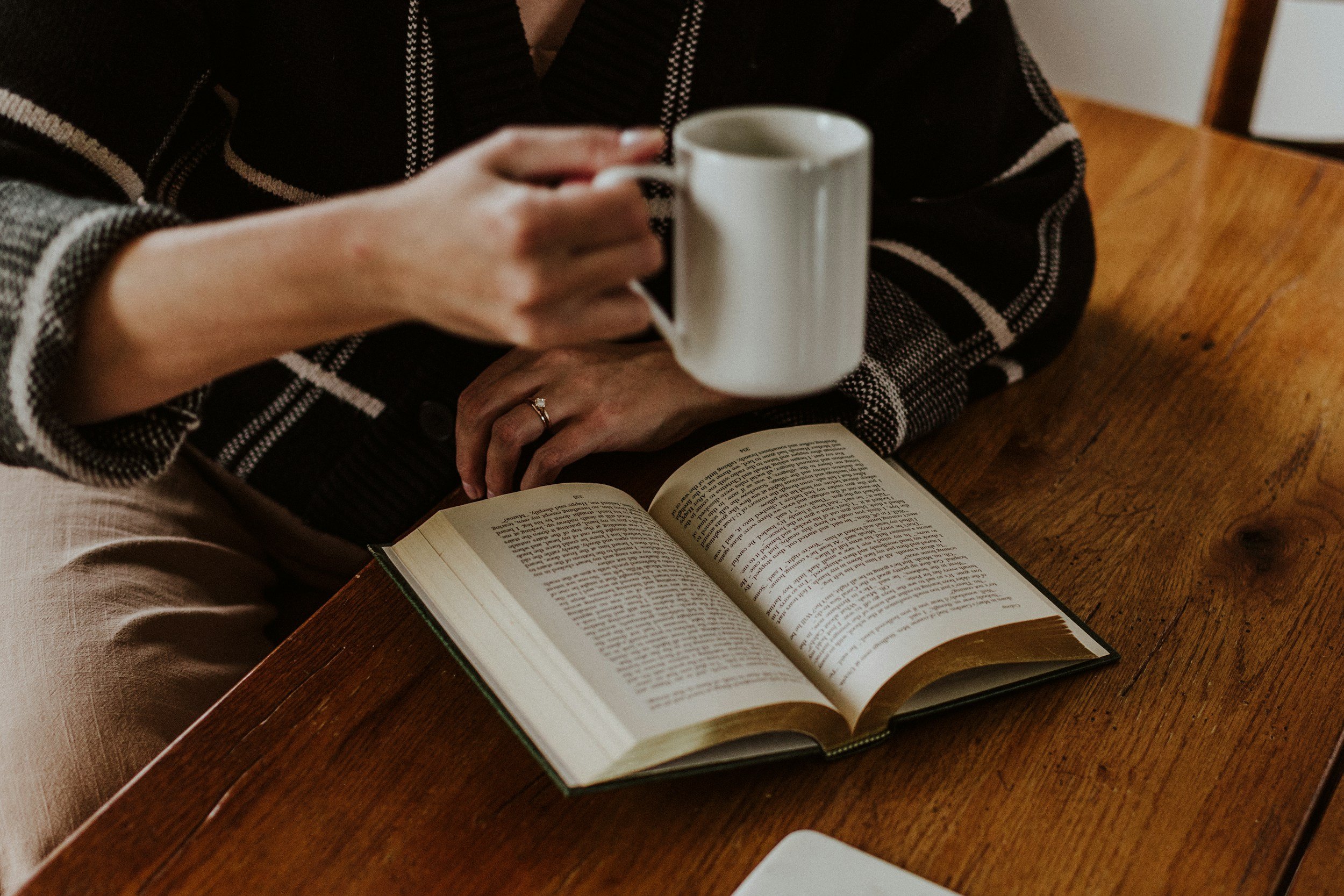 Person holding a white coffee mug with their right hand, resting on an open book on a wooden table.