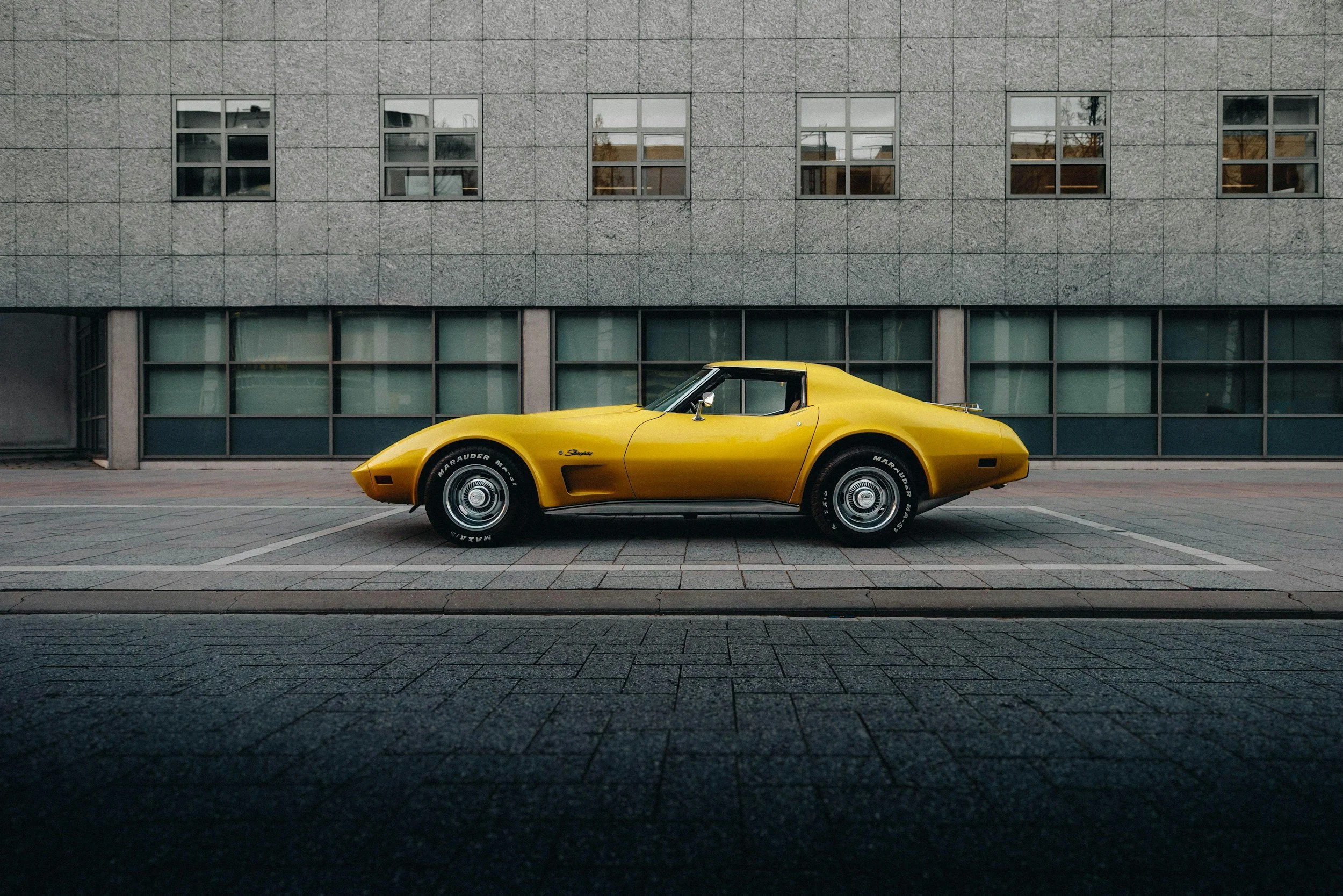 A classic yellow sports car parked on a city street in front of a modern concrete building with large windows.